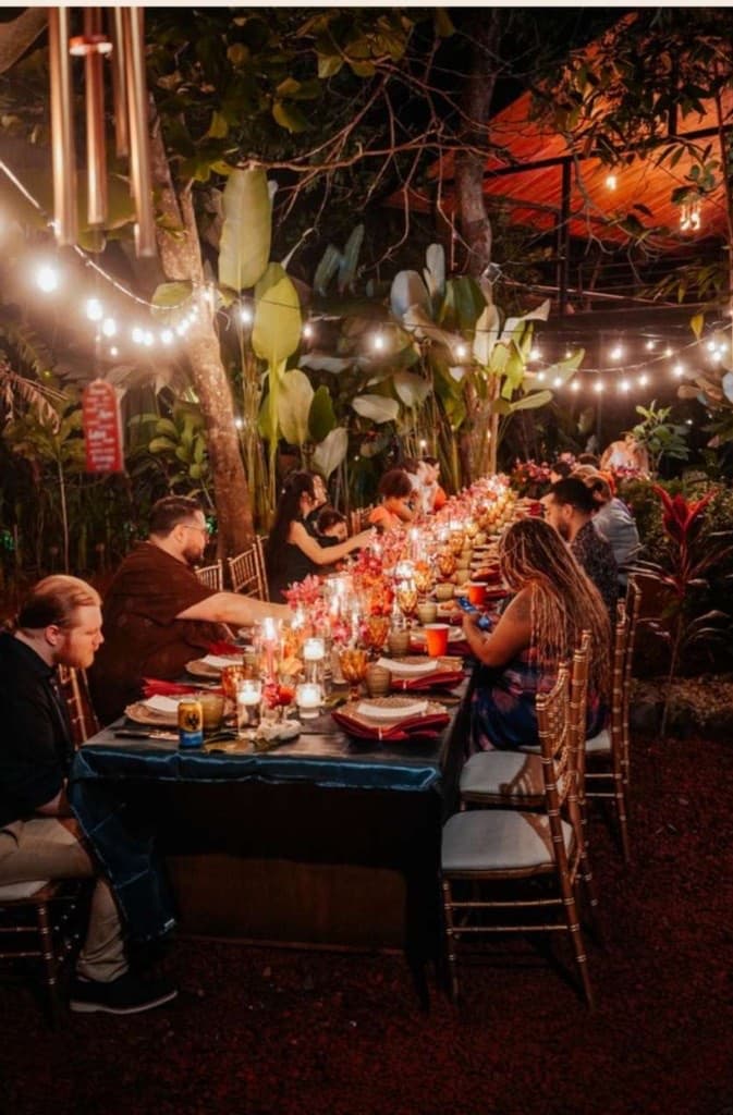Long candlelit communal dinner table under string lights with guests gathered in celebration